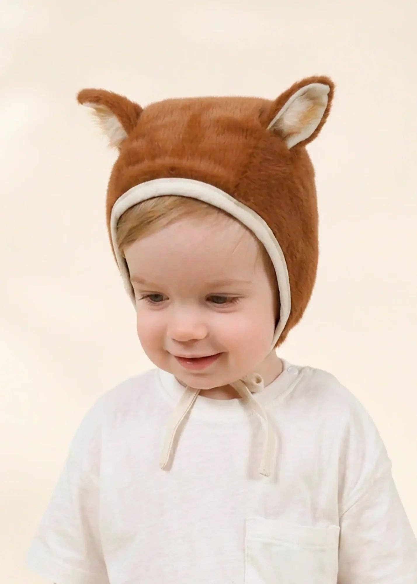 A toddler wearing a brown plush fox hood with long ears and a cream chin strap, paired with a white top, against a soft beige background.