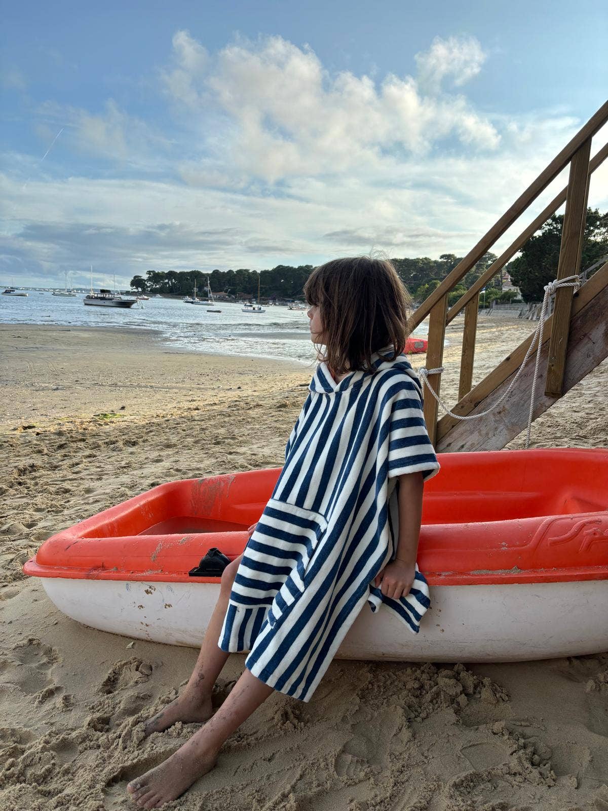 Person wearing a striped poncho, sitting on a red and white boat on a sandy beach with boats and trees in the background.