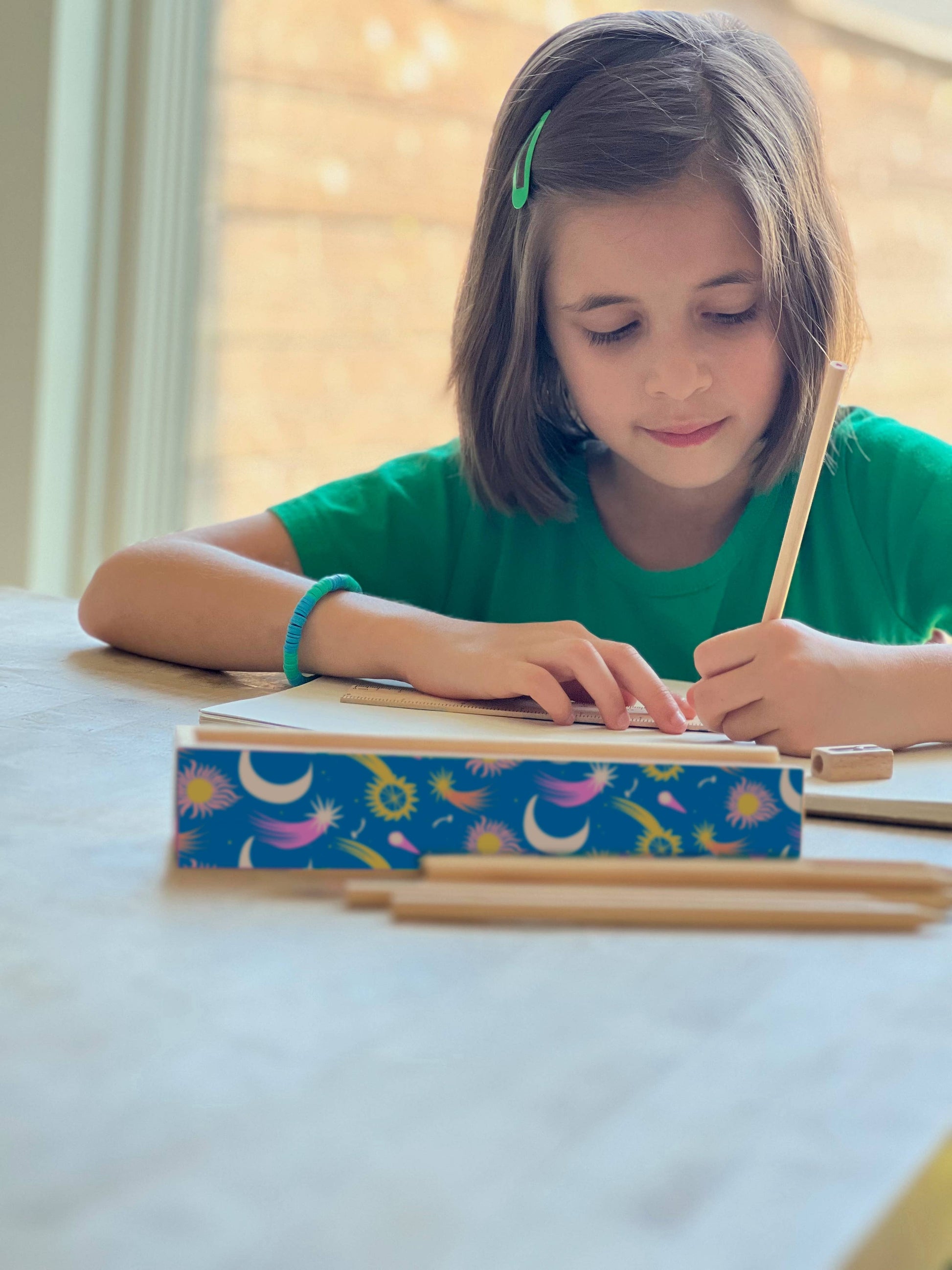 Young girl writing in a notebook with a colorful cover on a light surface.