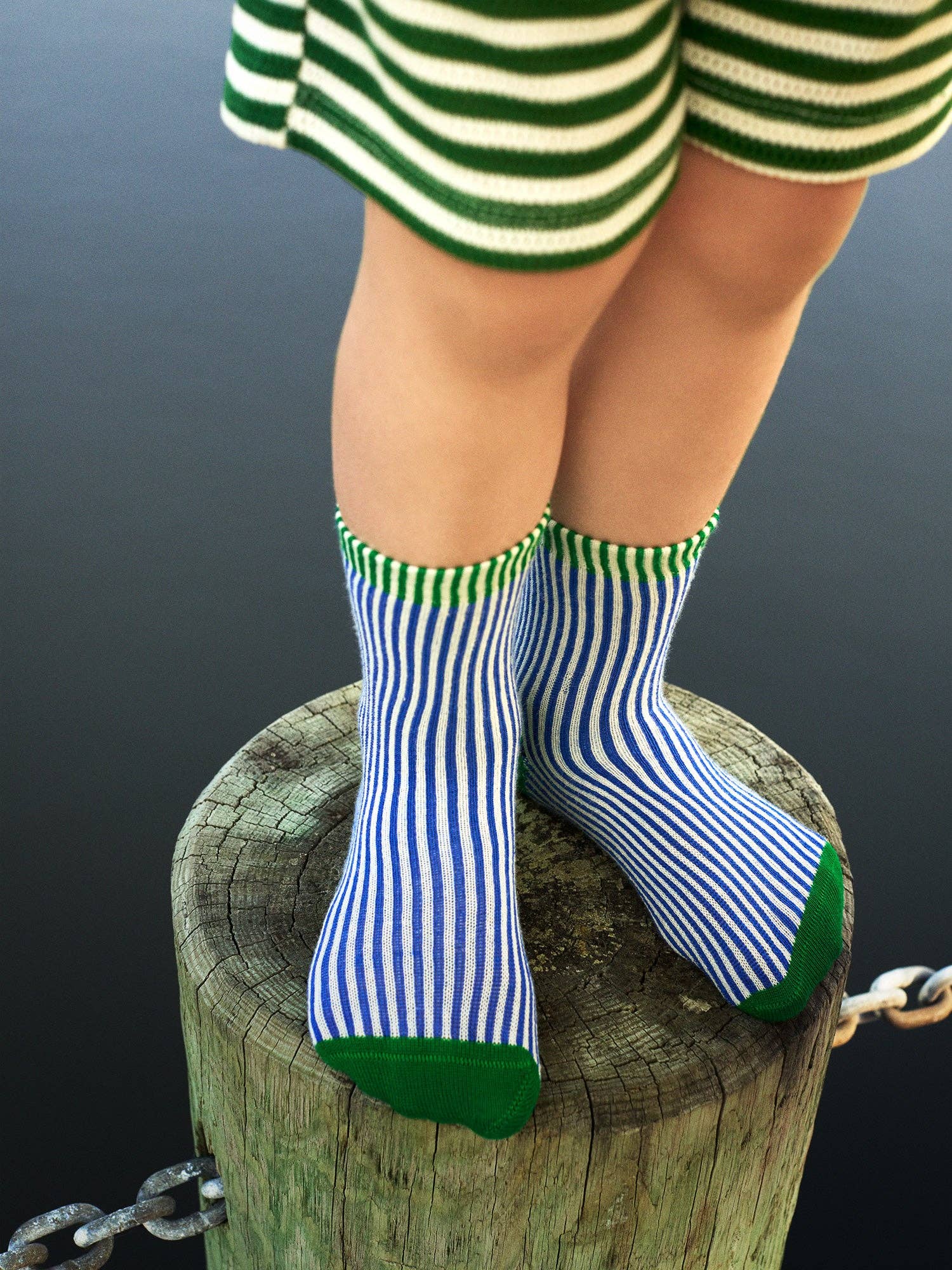 Child wearing green and white striped socks with blue and white striped pattern, standing on a wooden post against a dark background.