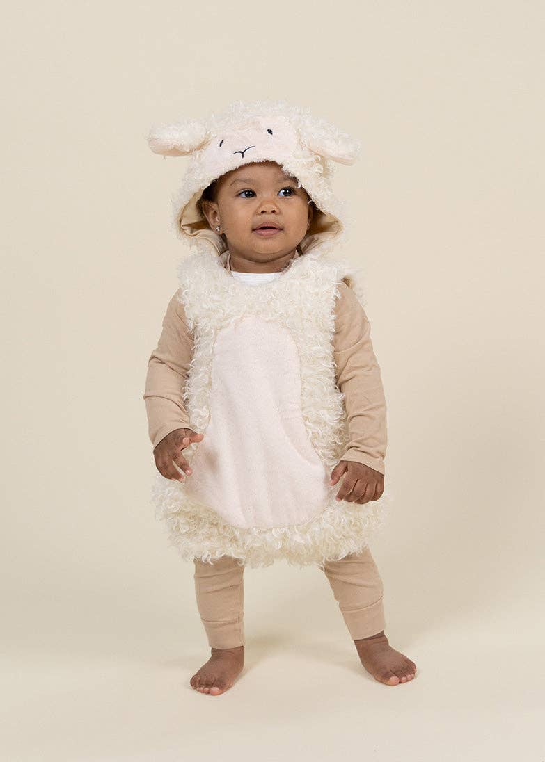 A toddler standing barefoot against a plain cream studio background, wearing a plush sheep costume with a hood featuring floppy ears, a fuzzy cream body, and beige leggings.