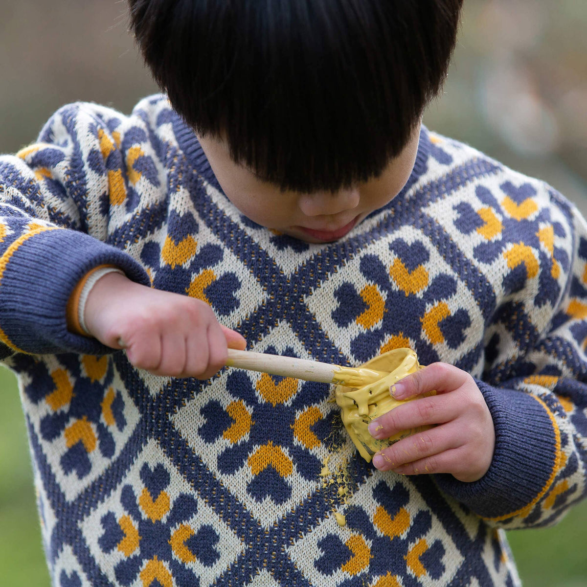 Child wearing a diamond-patterned knitted jumper while stirring yellow paint outdoors.