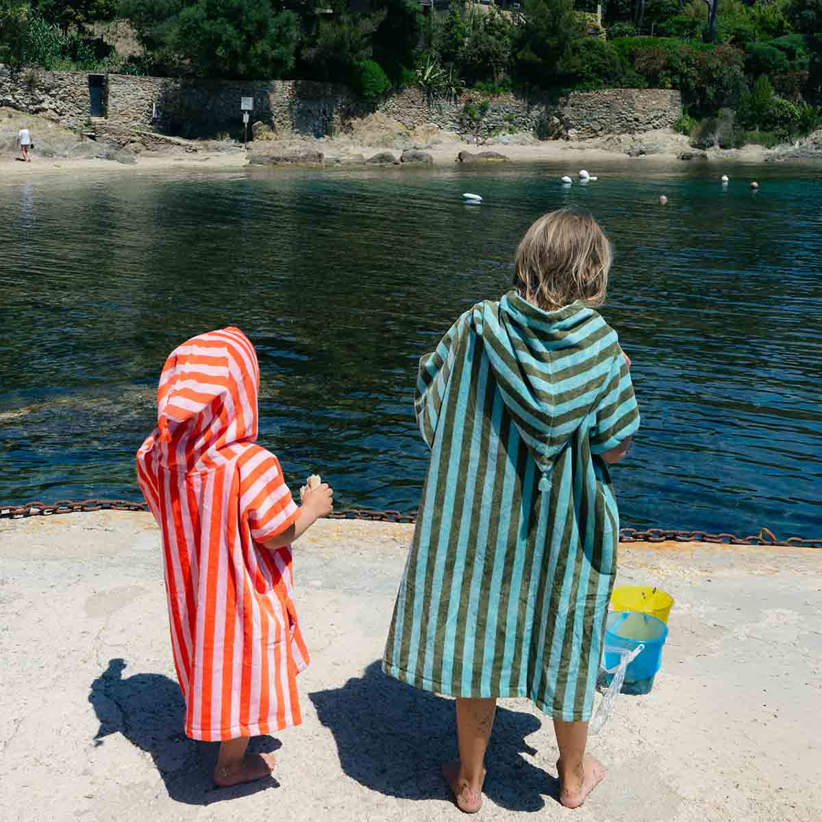 Two children in striped hooded towels standing by a body of water.