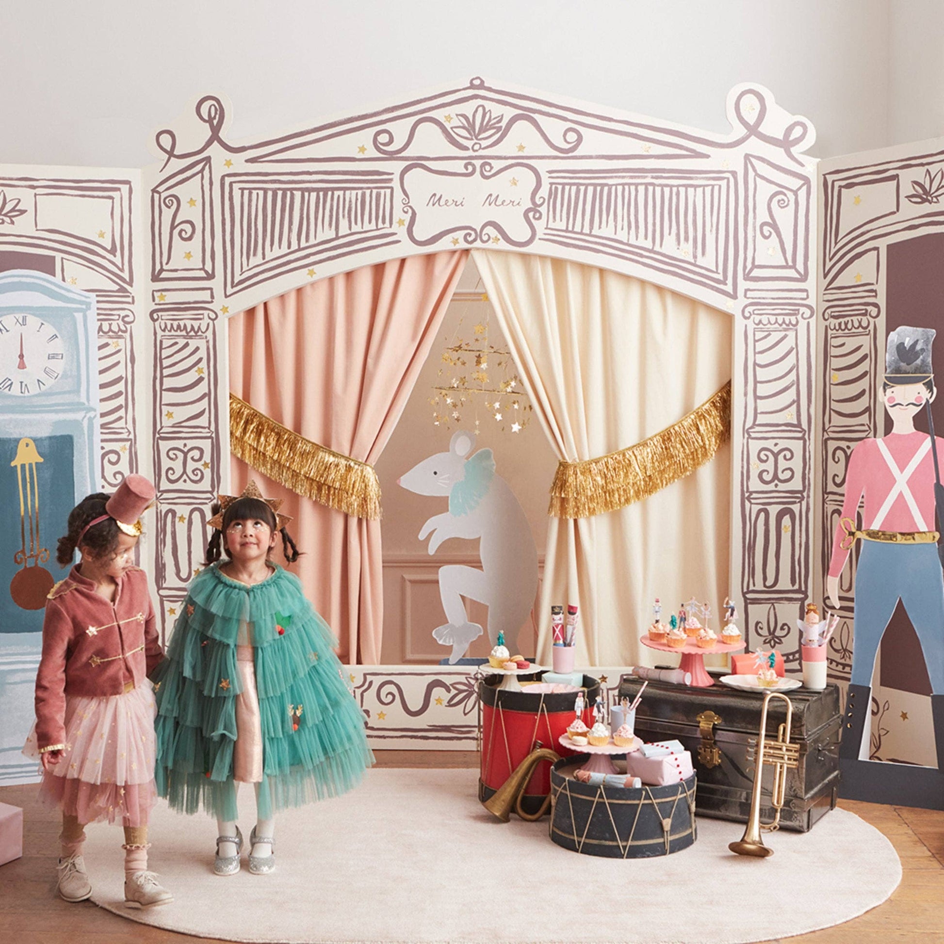 A children's playset or party backdrop featuring a vintage theater stage design with illustrated arches, curtains, a rabbit cutout, and props like drums, a trumpet, and a tea station. Two children are standing in front of the set on a round rug in a bright indoor room.