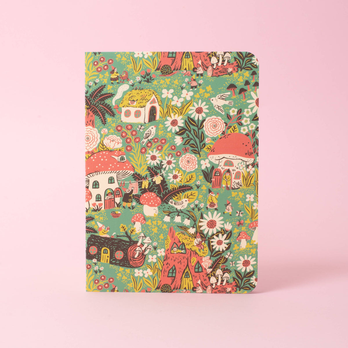 A softcover saddle-stitch notebook with vibrant wraparound cover featuring toadstool houses and flowers and gnomes, shown on a pink background