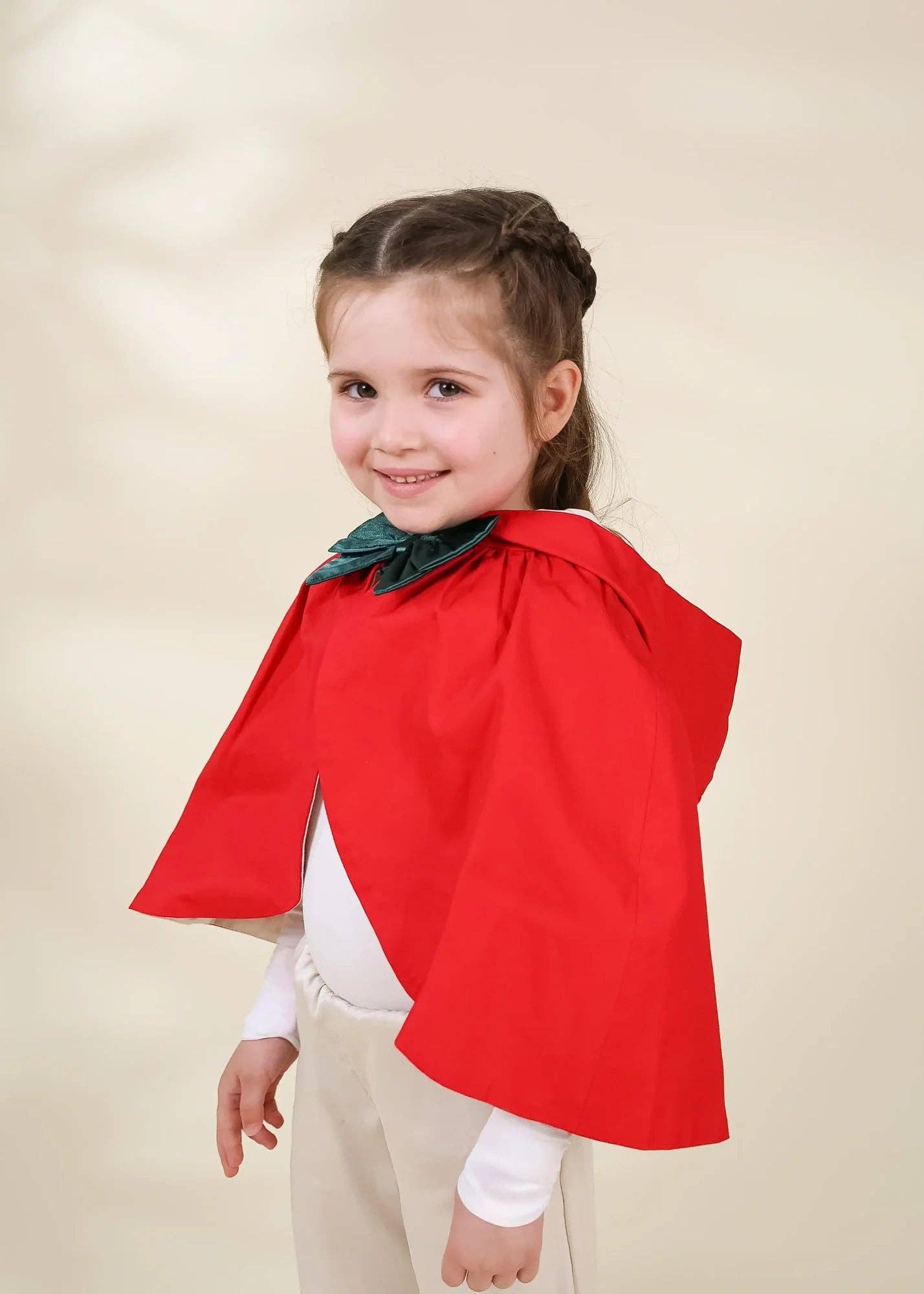 A young girl with her hair in a half-up braid wearing a bright red cape with a dark green bow accent, a white long-sleeve shirt, and beige pants, set against a soft beige studio background.