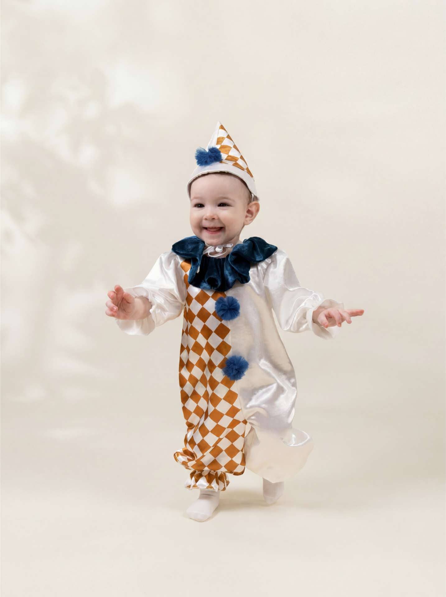 A baby wearing a white and gold checkered clown costume with a navy blue bow tie and matching pointed hat, smiling and posing against a soft beige background.