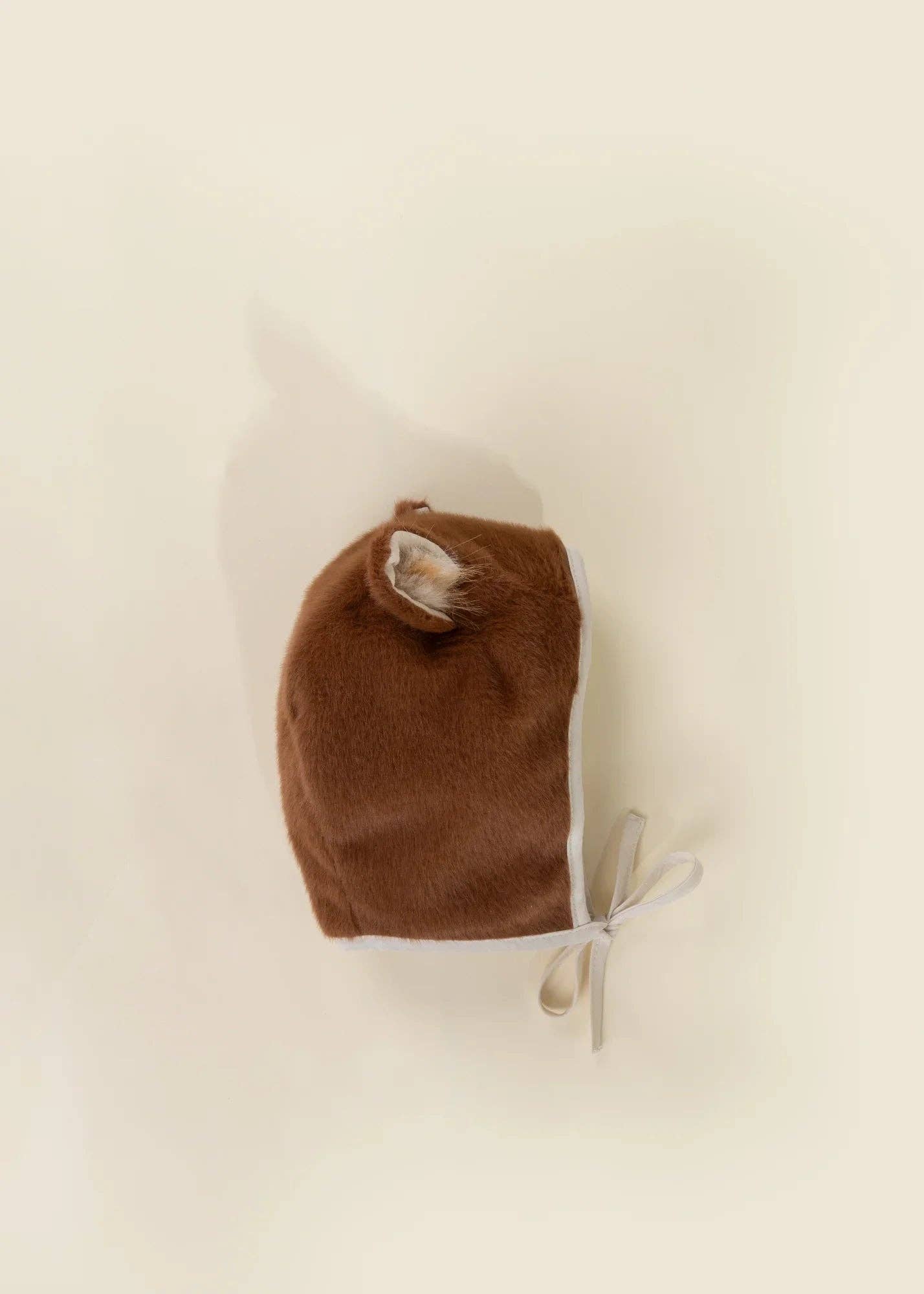 A brown plush bonnet with a small faux fur ear detail and a white ribbon tie, photographed flat on a warm cream background.