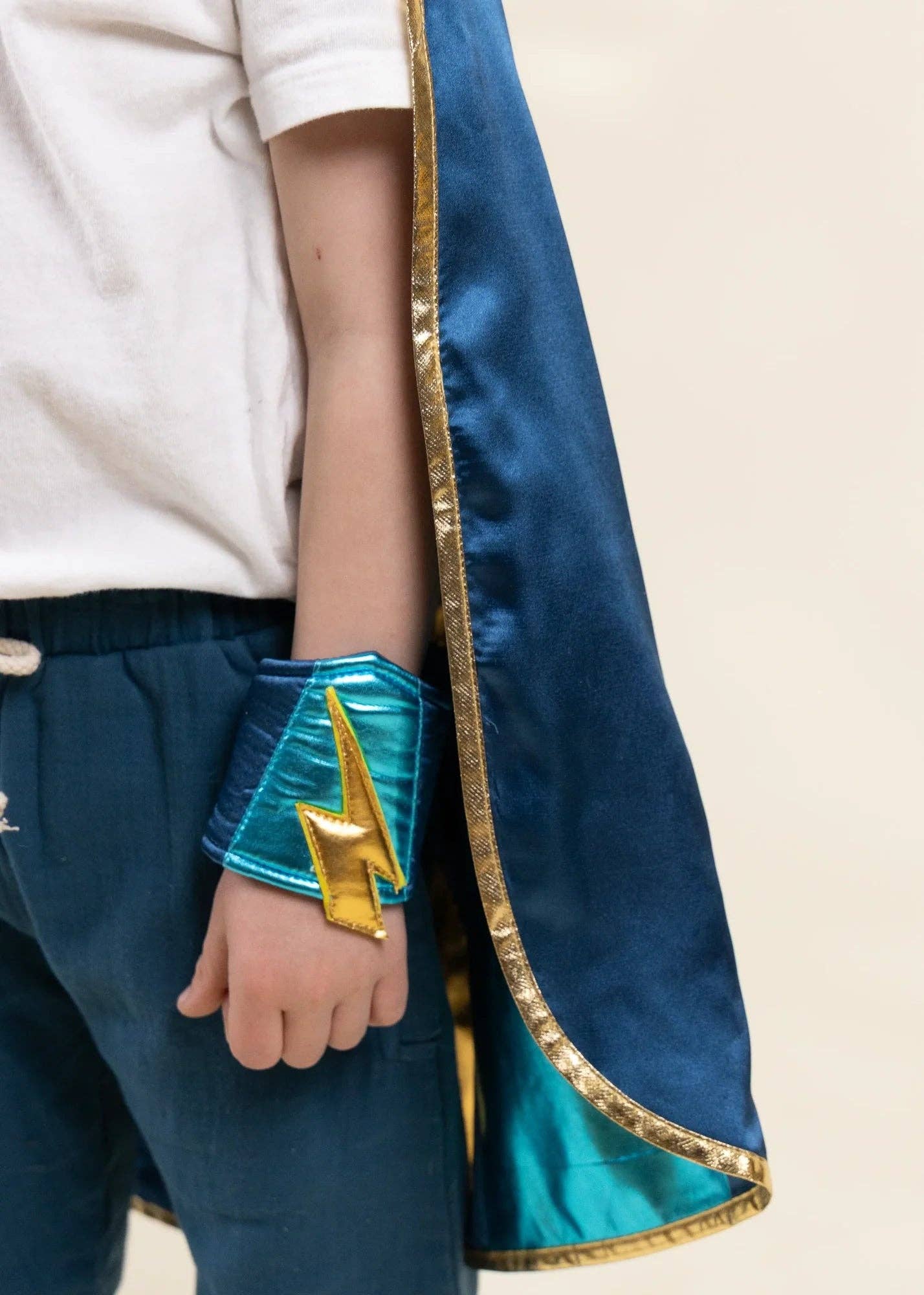 A close-up of a child wearing a blue superhero cape with gold trim, a matching blue wrist cuff, and a gold lightning bolt accessory, paired with a white t-shirt and dark pants.
