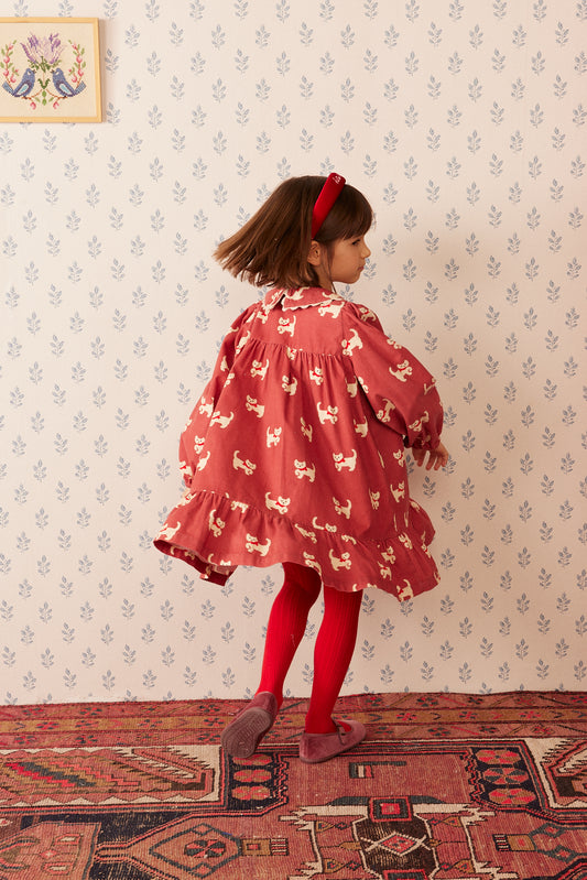 A young girl is shown from the back wearing a dusty red tiered dress with a white dog print, red tights, and a red headband, standing on a patterned rug against a floral wallpaper background.