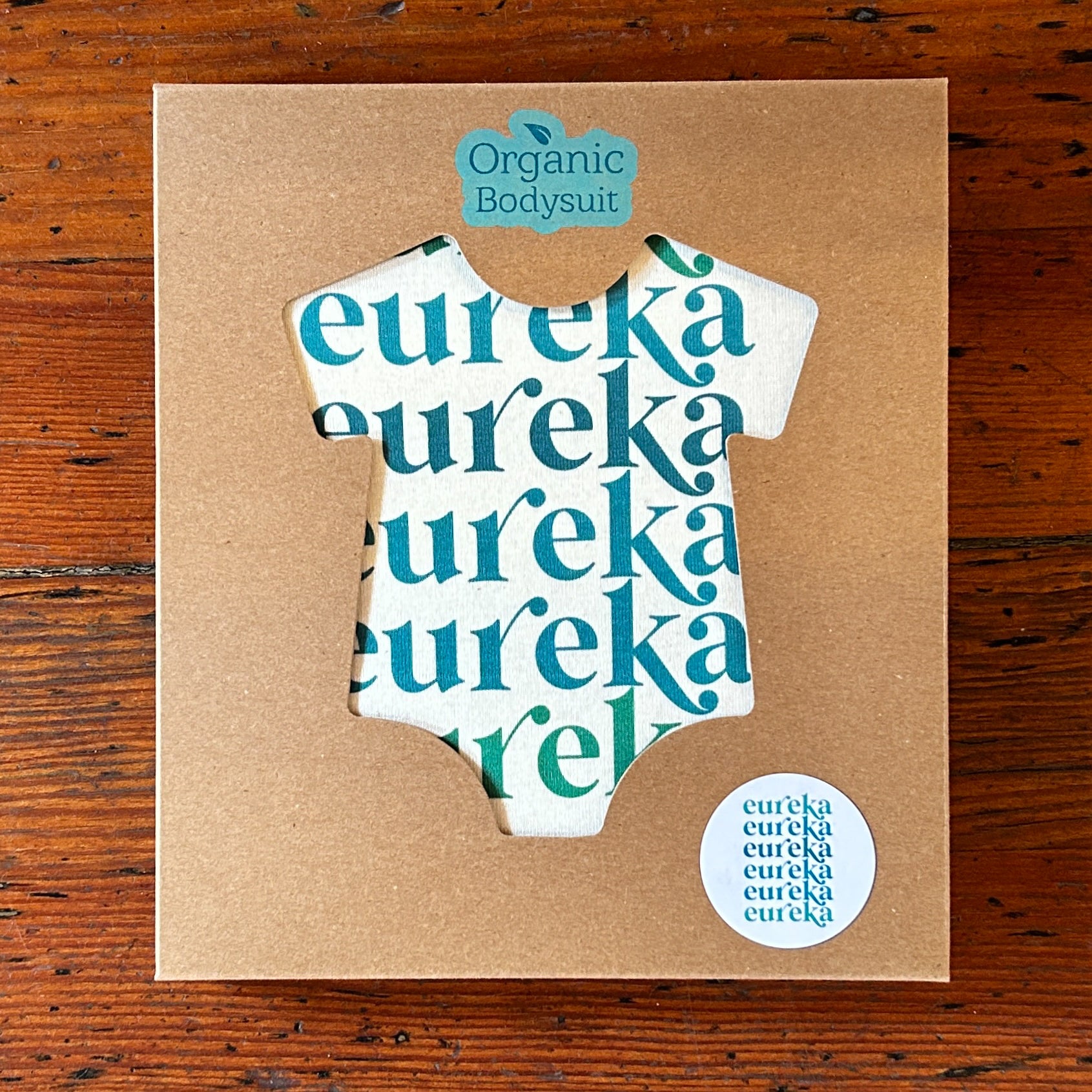 Cardboard packaging with a cut-out shape of an organic bodysuit, with onesie inside featuring the word 'eureka' repeatedly.