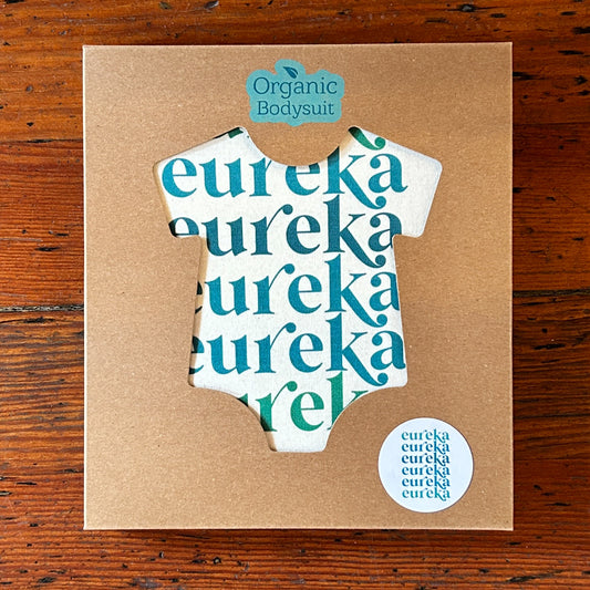 Cardboard packaging with a cut-out shape of an organic bodysuit, with onesie inside featuring the word 'eureka' repeatedly.