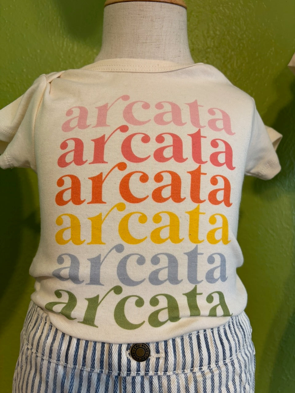 children's cream colored onesie with "arcata" repeated on the chest in various colors, on a mannequin, with striped pants