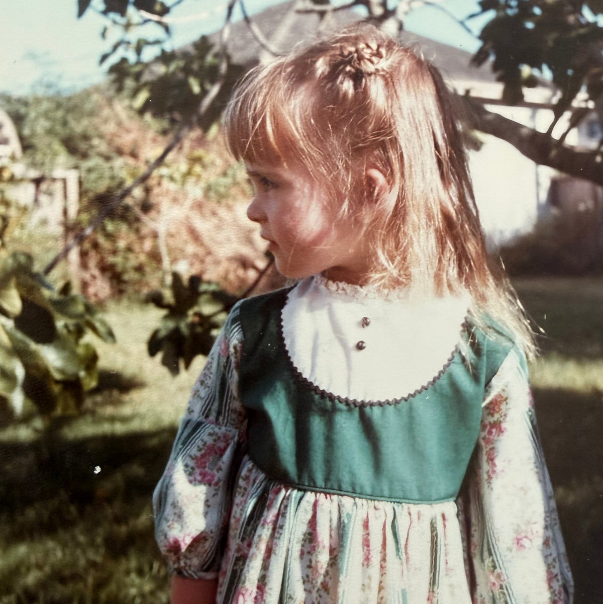 A young girl wearing a vintage-style floral dress with a solid green bodice and a large white collar, standing outdoors under a tree.