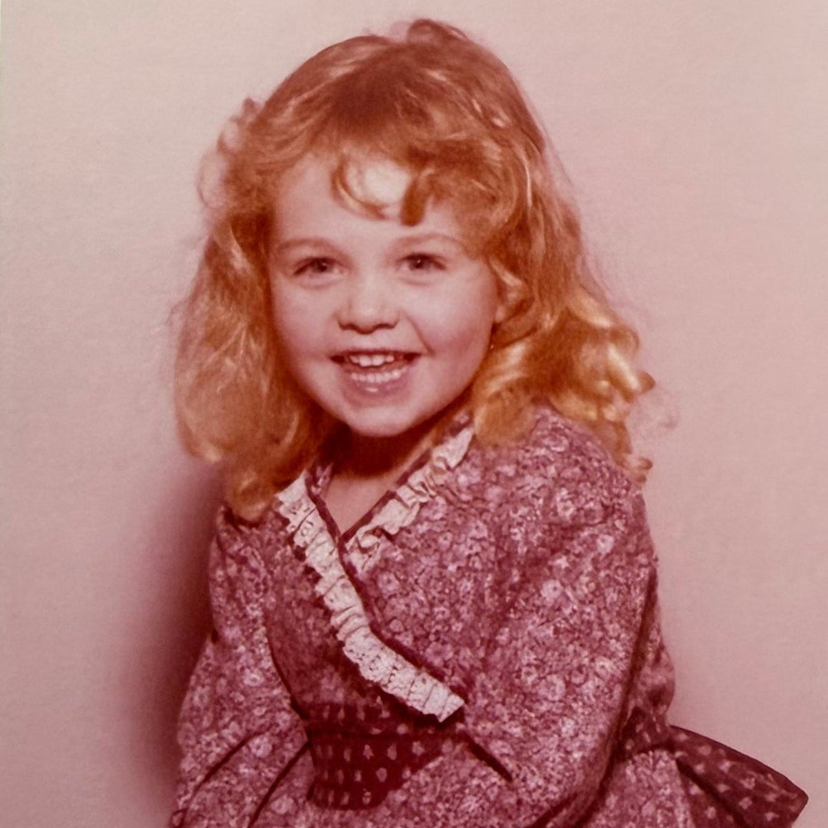 A vintage color photograph of a young girl with blonde curly hair, smiling and sitting cross-legged. She is wearing a long-sleeved, dark reddish-brown floral patterned dress with a white ruffled collar.