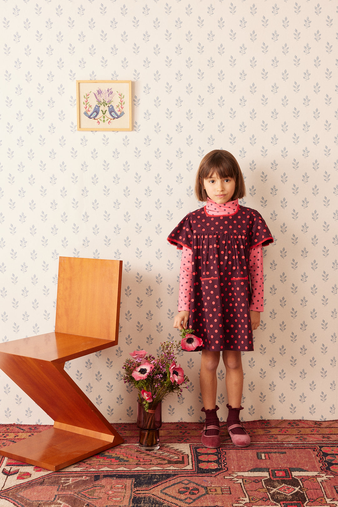 Girl wearing Mariana plum dress by Hello Simone standing in wallpapered room next to modern chair and bouquet of flowers