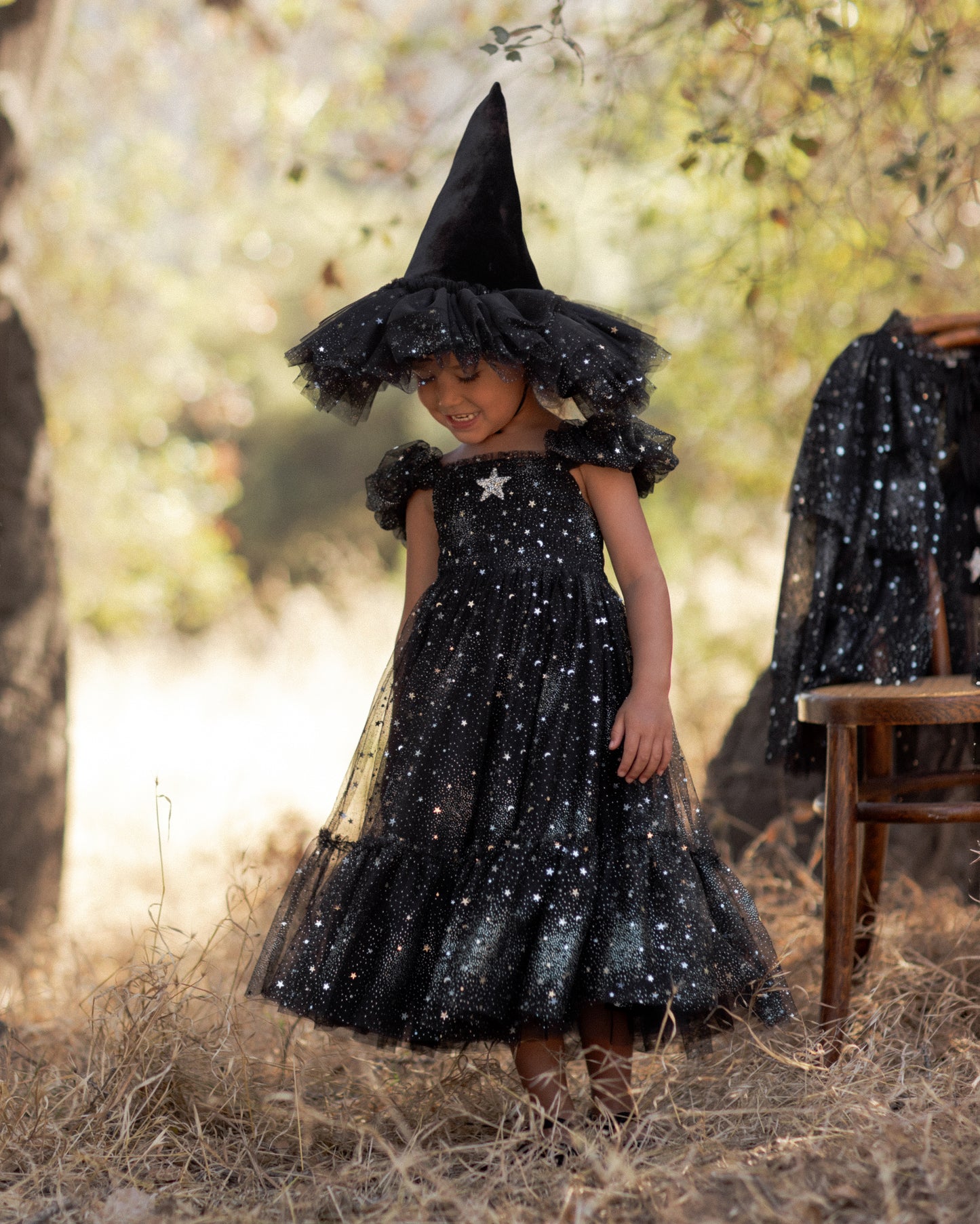Young child smiling, wearing a black dress with silver starts and a matching witch hat standing in a field next to a tree