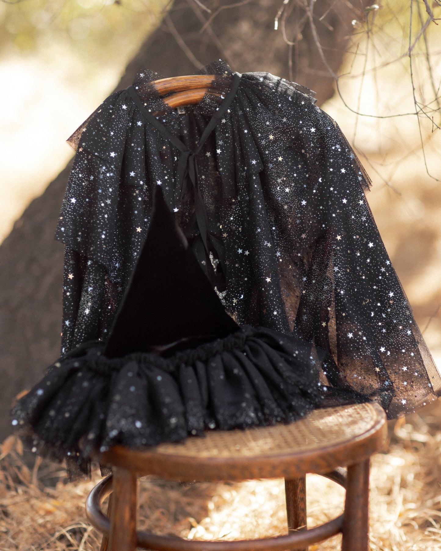 Black tulle cape for children with silver starts on it draped over a wooden chair in front of a tree trunk