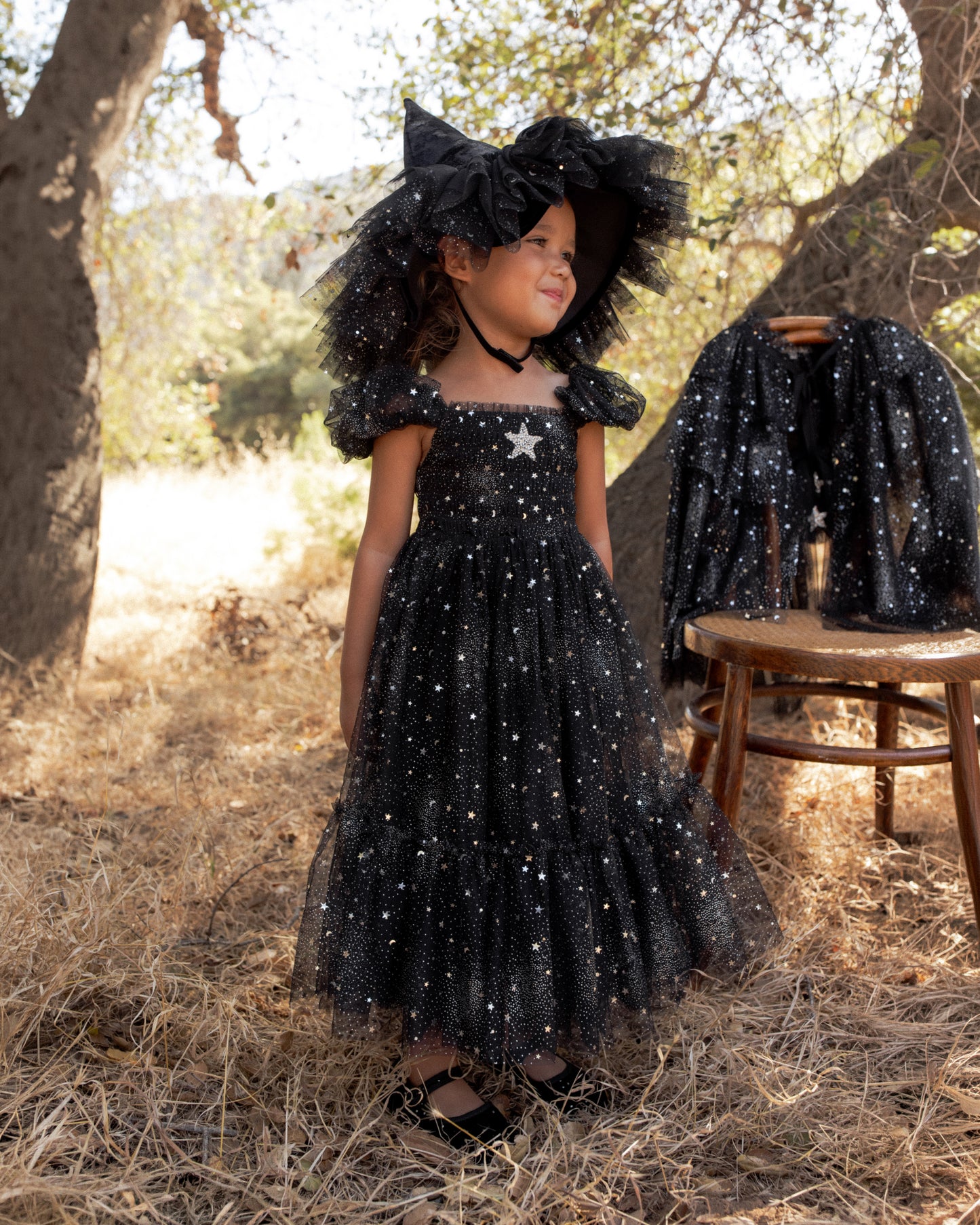 Young child wearing a black dress with silver starts and a matching witch hat standing in a field next to a tree