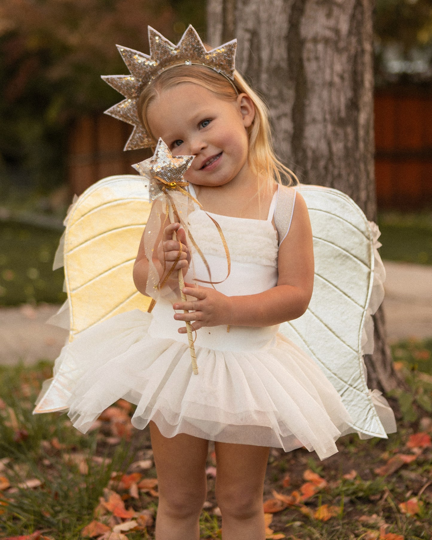 young child wearing an angel costume wearing a gold sun crown and holding a gold star wand standing in front of a tree