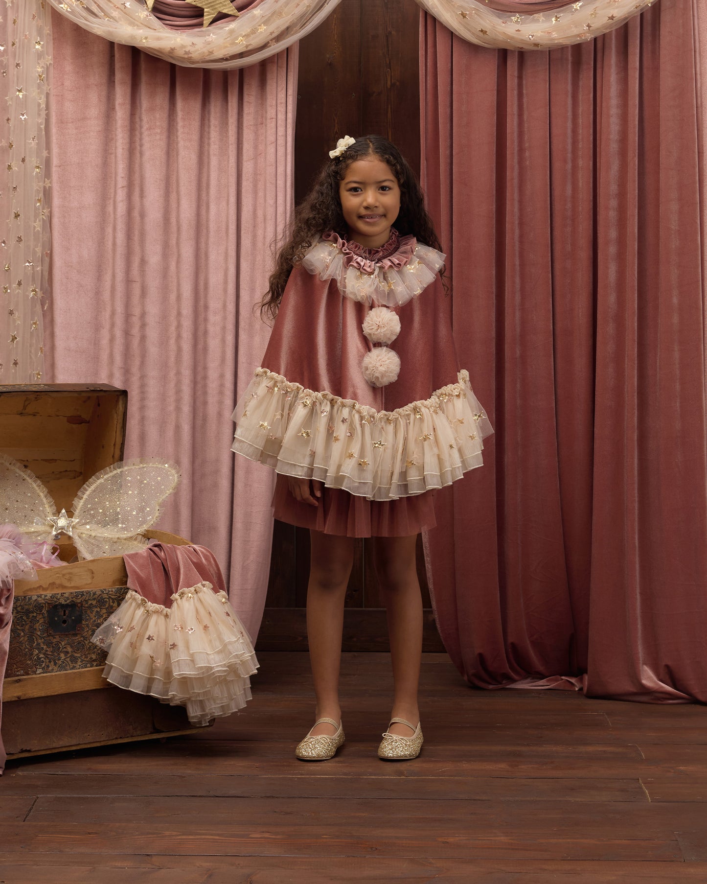 Child wearing a dark pink clown cape with ivory ruffles and pom poms, standing in front of dark pink curtains