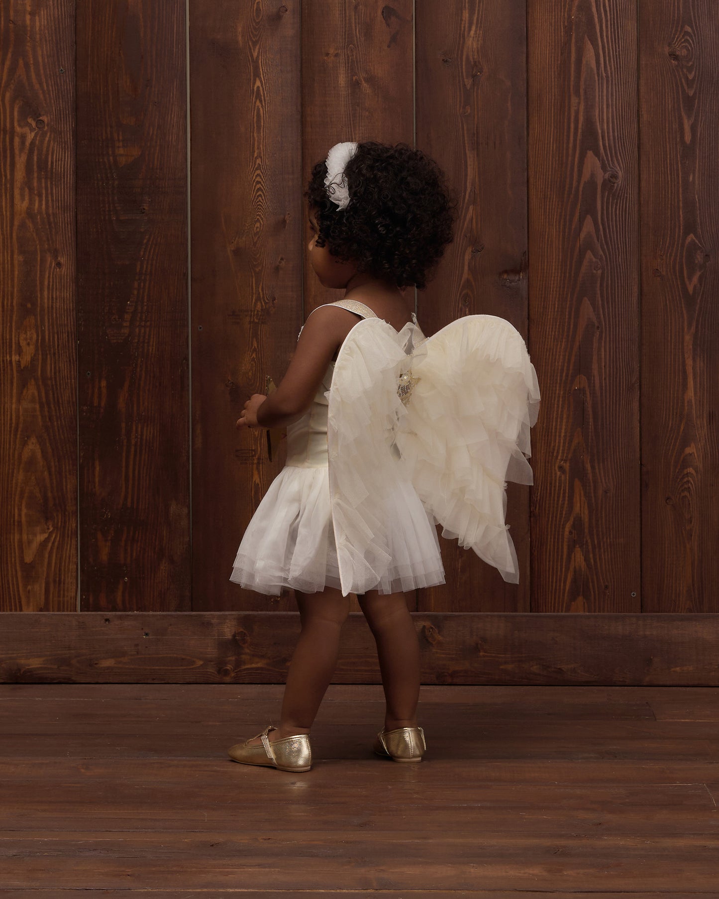 Young child wearing angel wings and a white dress, a white headband, and gold shoes, standing in front of a wooden background