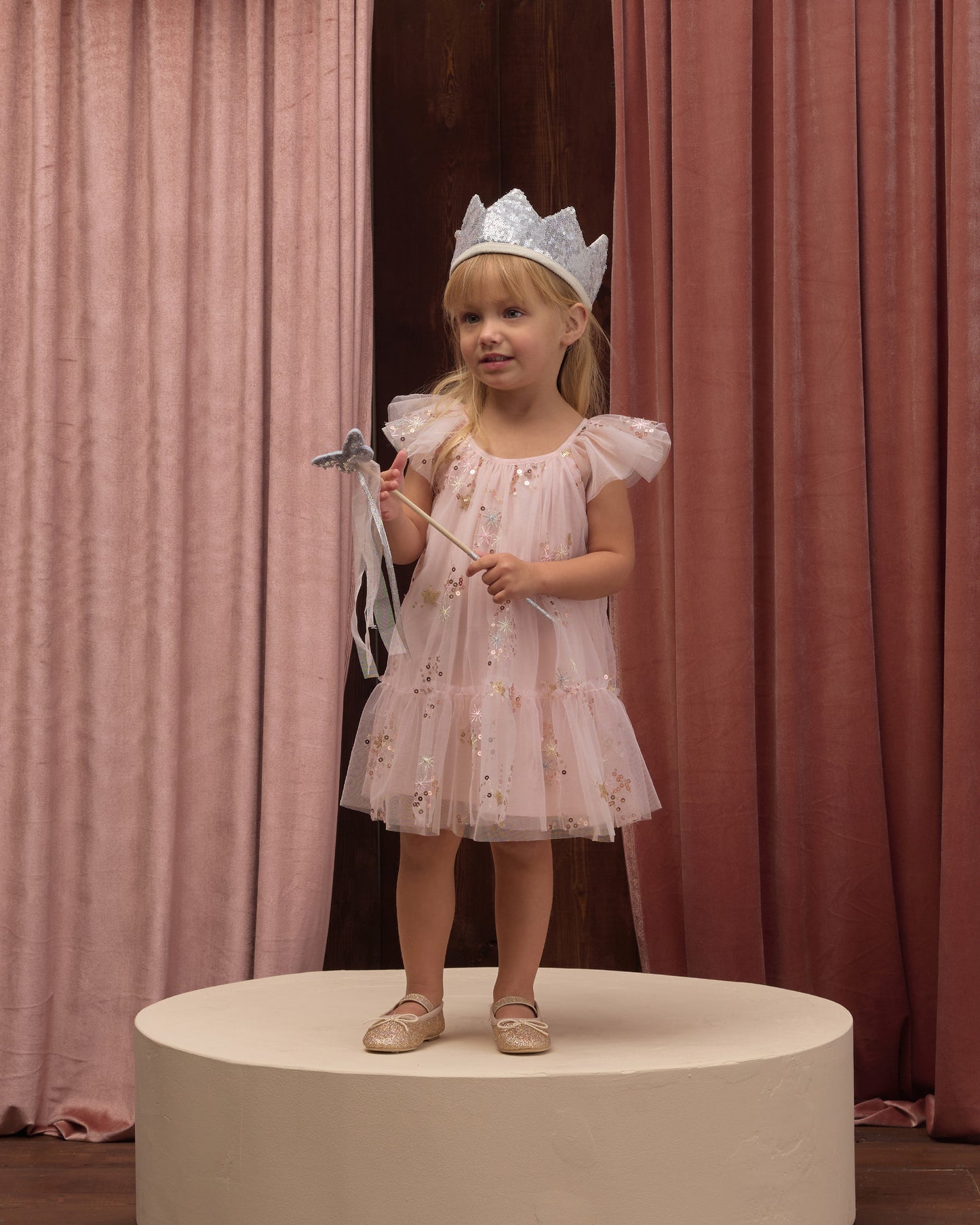 Young child wearing a pink dress and a silver sequin crown, holding a silver sequin wand