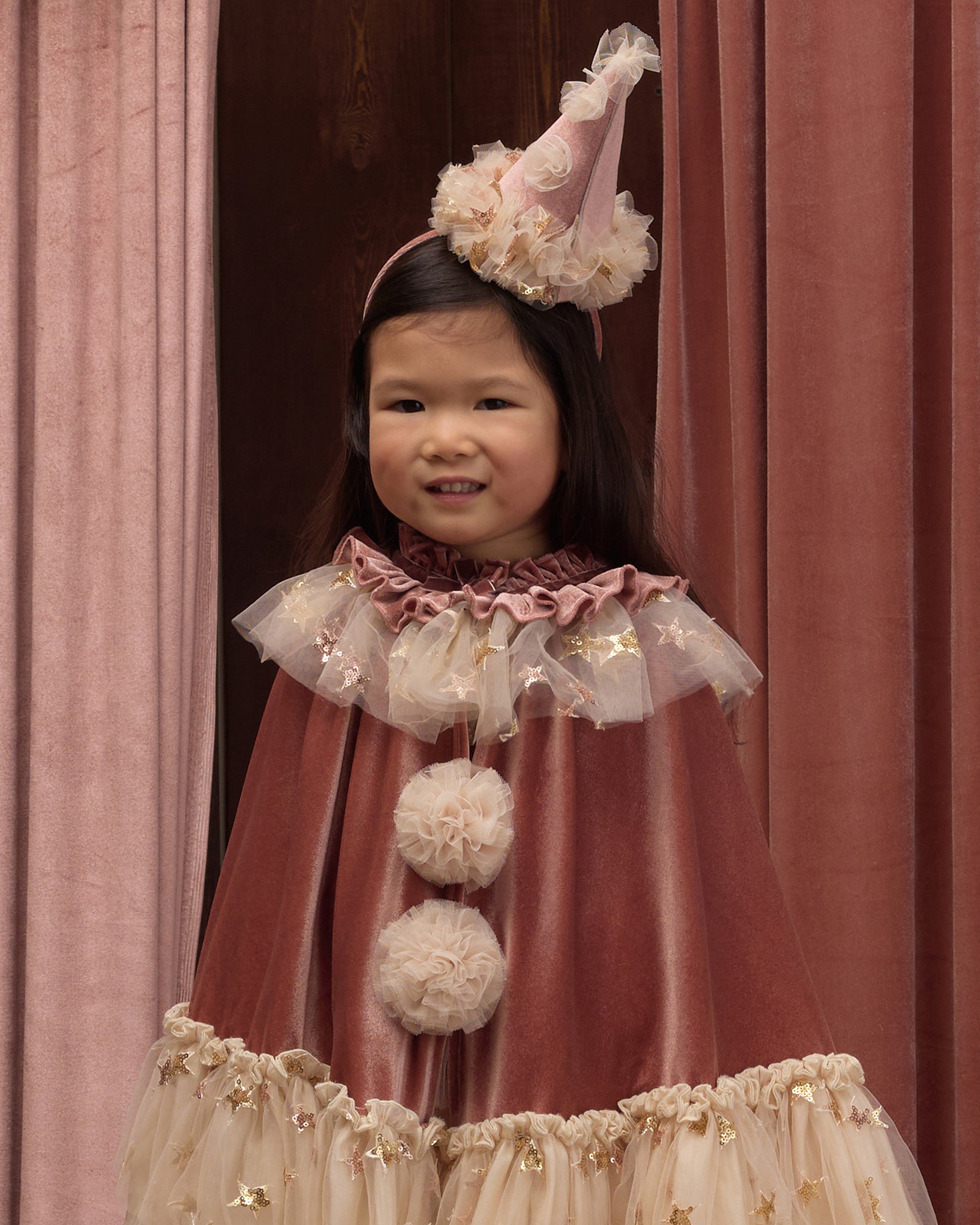 Young girl wearing a clown cape and matching clown hat in front of drapes