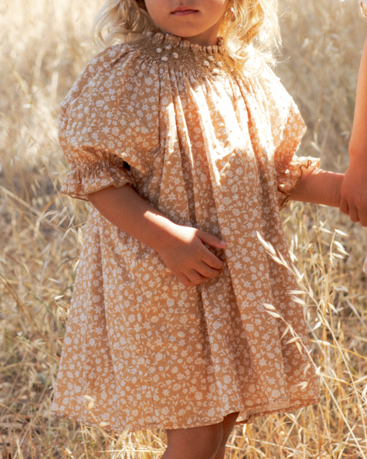 A young child wearing a short puff-sleeve dress with a small white floral print on a warm tan background, standing outdoors in a field of dry golden grass.