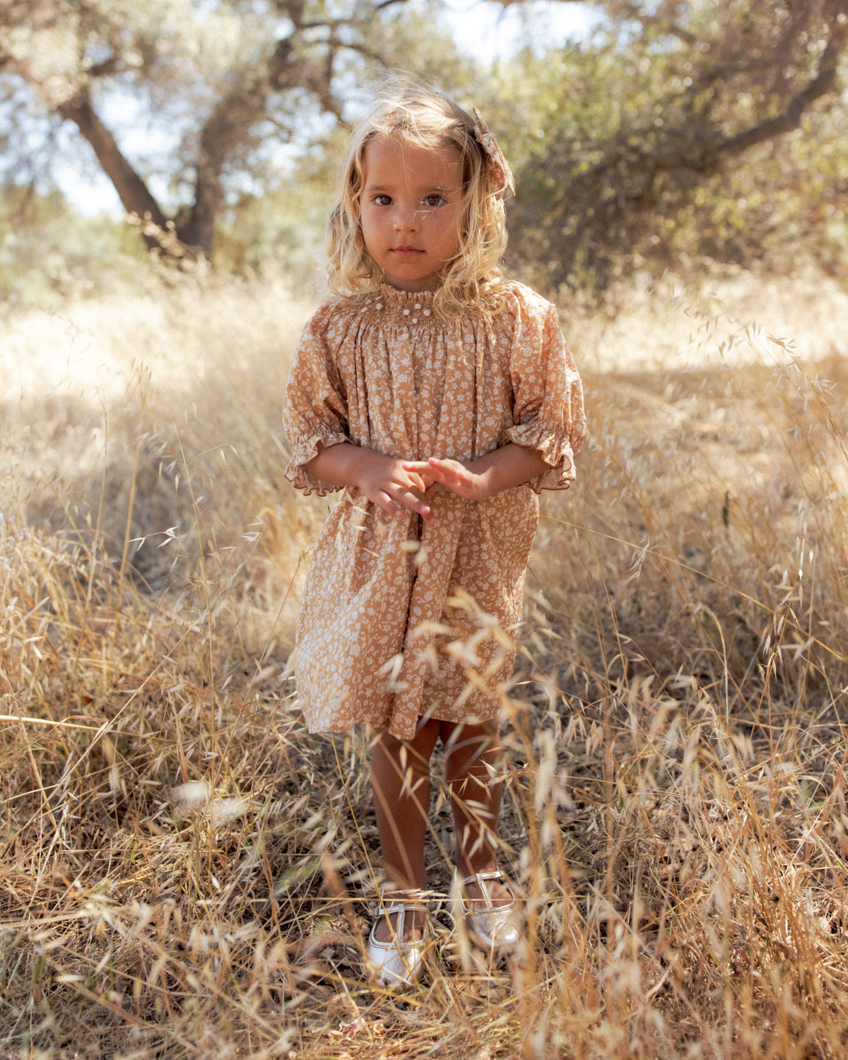 A young girl with blonde hair stands in a dry grassy field wearing a beige and brown patterned dress with ruffled sleeves and silver shoes.