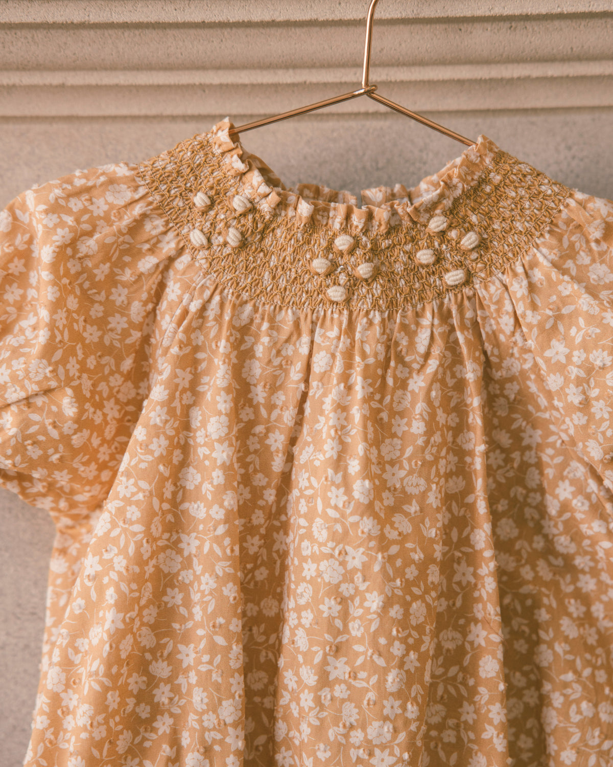 A close-up of a short-sleeve blouse hanging on a gold hanger against a textured beige wall. The blouse features a warm mustard yellow base with a delicate white floral pattern and a smocked yoke decorated with small white pom-poms.