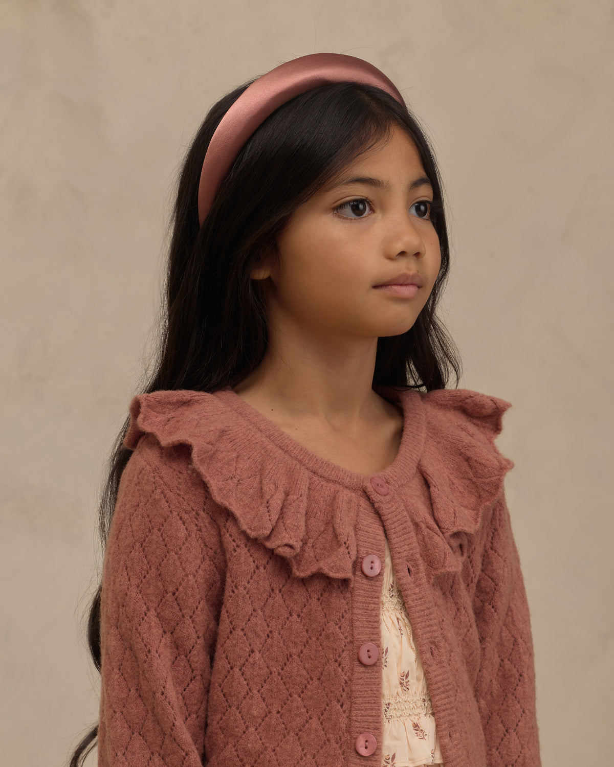 A young girl with long dark hair wearing a dusty rose pink ribbed knit cardigan with ruffle details and a matching pink satin headband, set against a neutral textured beige background.