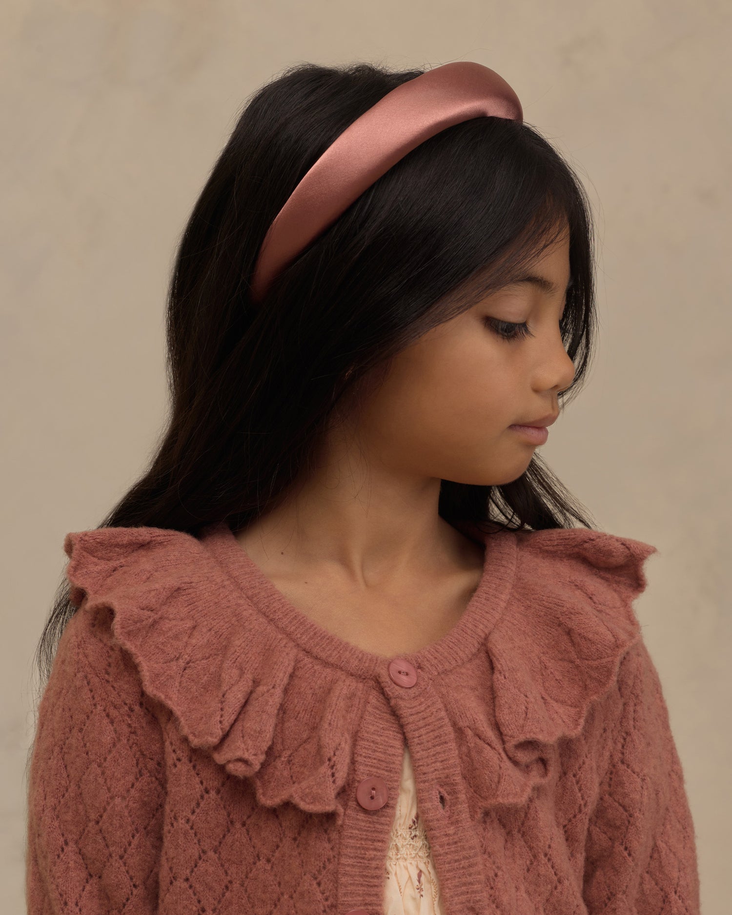 A young girl with long black hair wearing a dusty rose satin headband and a matching textured knit cardigan with a ruffled collar, shown in profile against a neutral beige background.