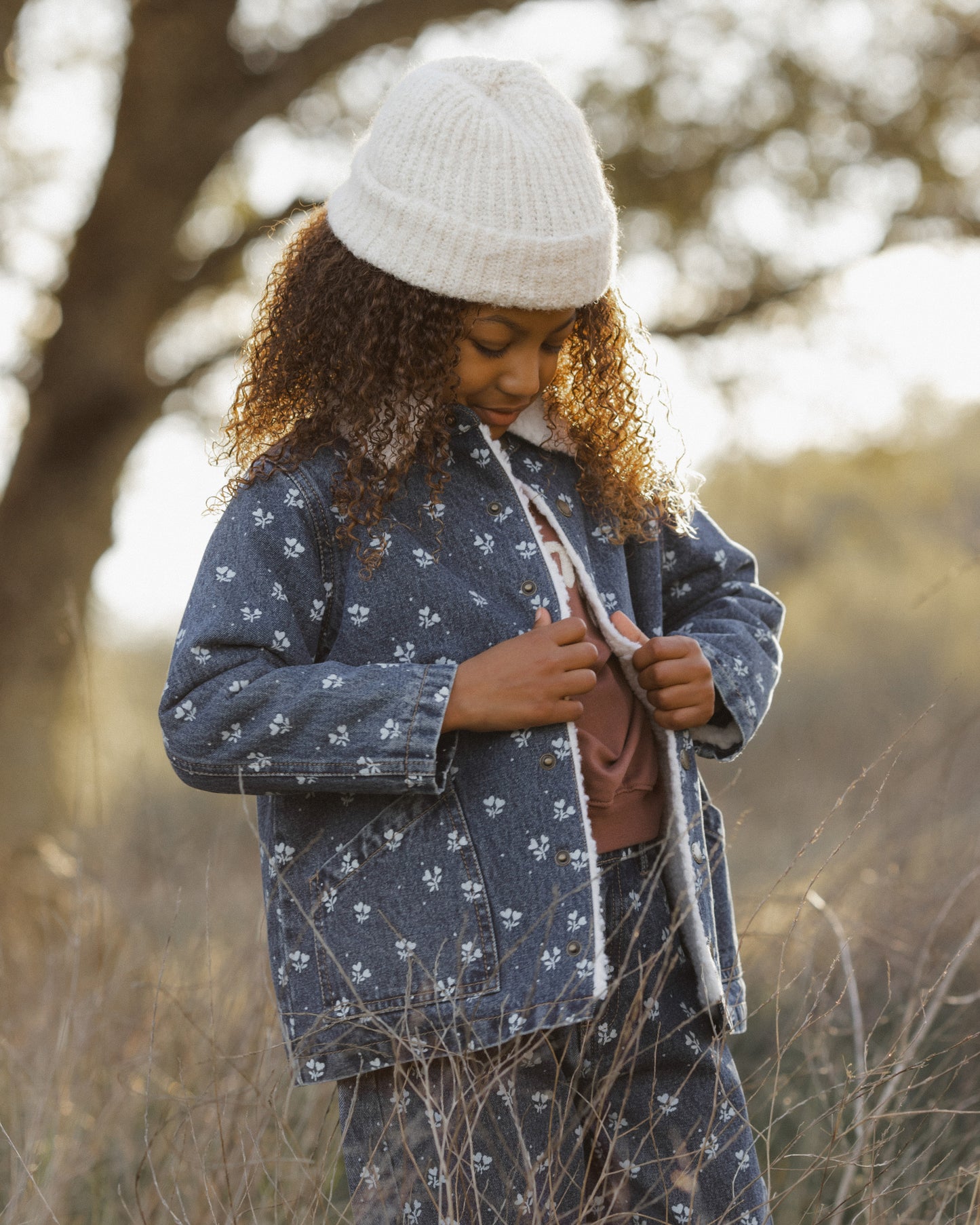 A child wearing a matching navy blue coat and jeans set with a white floral print,  styled with a white knit beanie in an outdoor field.