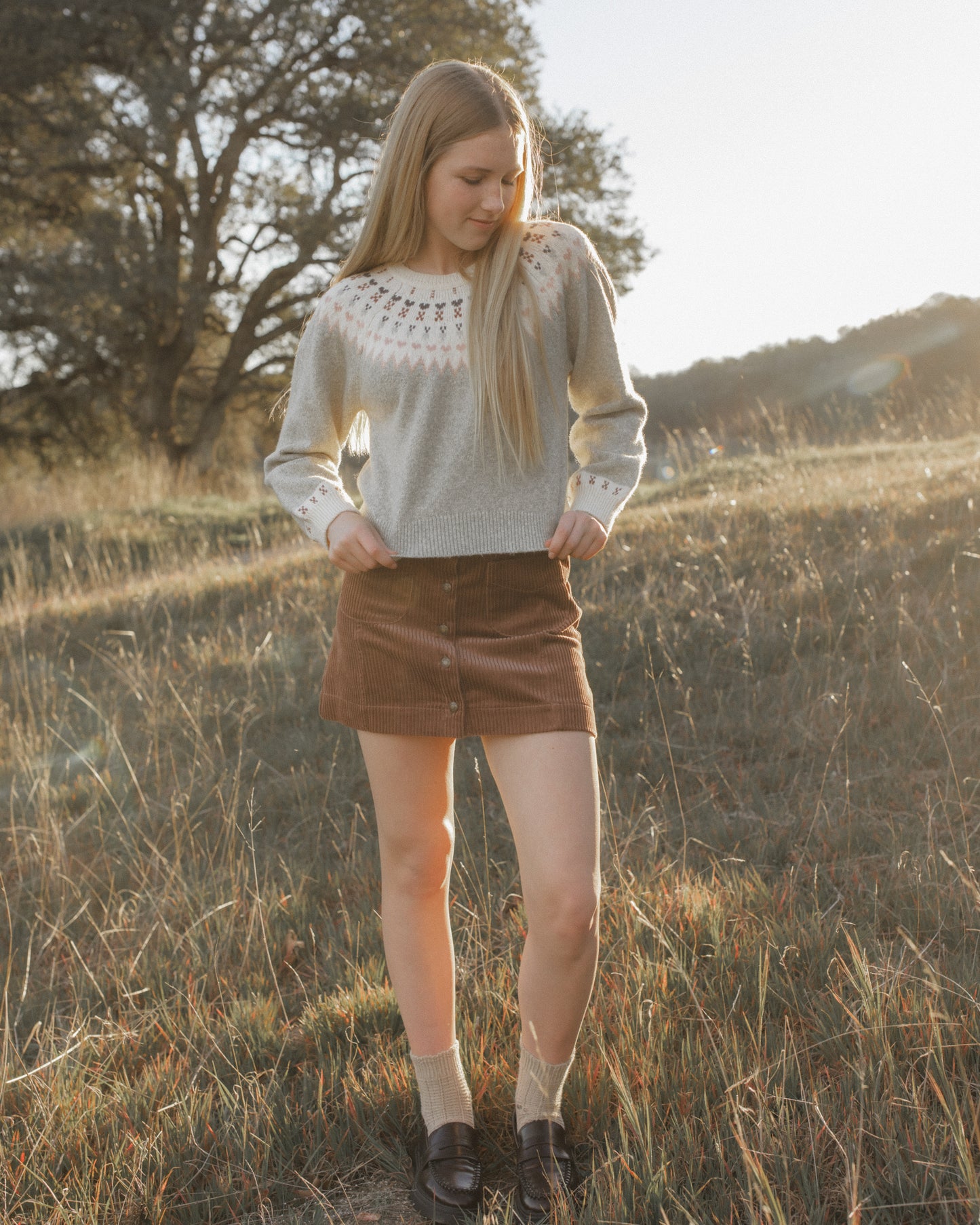 A young woman stands in a sunlit grassy field wearing a light gray knit sweater with a white crochet yoke, a brown mini skirt, beige socks, and dark loafers.