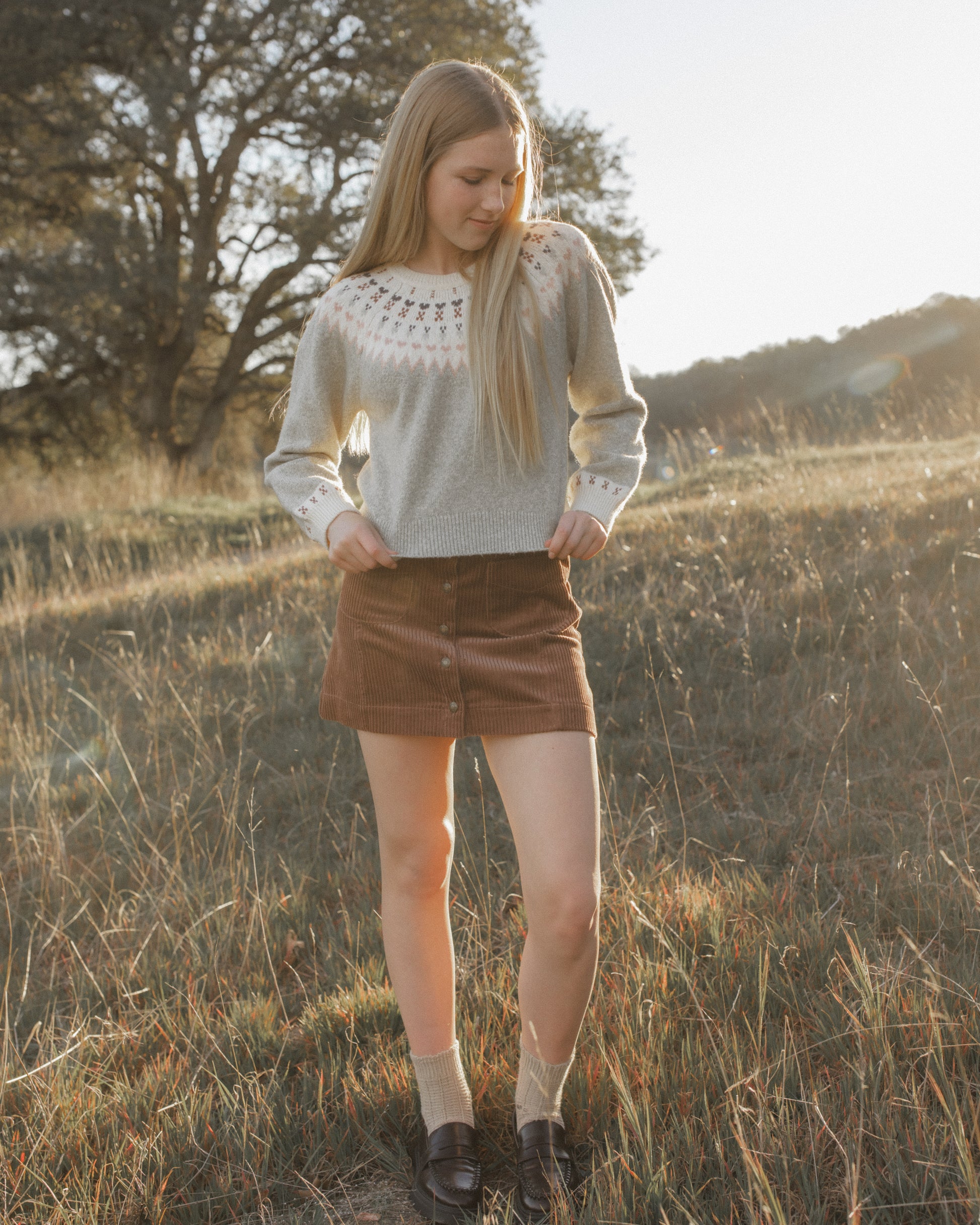 A young woman stands in a sunlit grassy field wearing a light gray knit sweater with a white crochet yoke, a brown mini skirt, beige socks, and dark loafers.