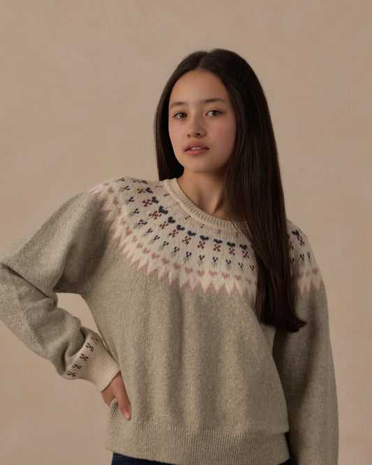 A woman wearing a light beige knit sweater with a Fair Isle patterned yoke in cream, pink, and dark navy, posed against a solid beige background.