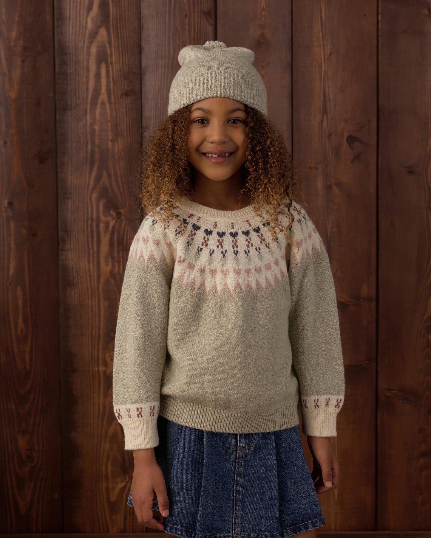 A young girl wearing a beige knit sweater with a pink and dark blue Fair Isle yoke pattern, a matching beige knit beanie, and a blue denim skirt, standing against a dark wood plank wall.