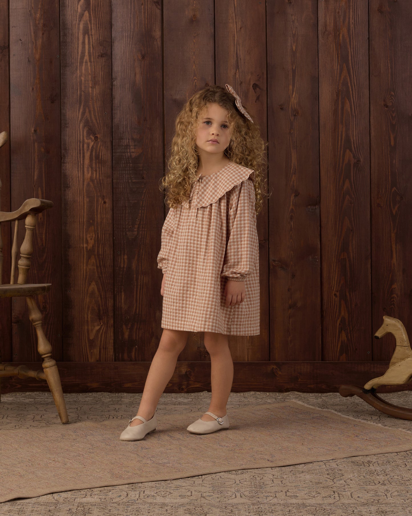 A young girl with curly light brown hair stands in front of a dark wood plank wall wearing a beige and white gingham dress with a large ruffled collar and white flats. A wooden chair is partially visible on the left, and a rocking horse is partially visible on the right.