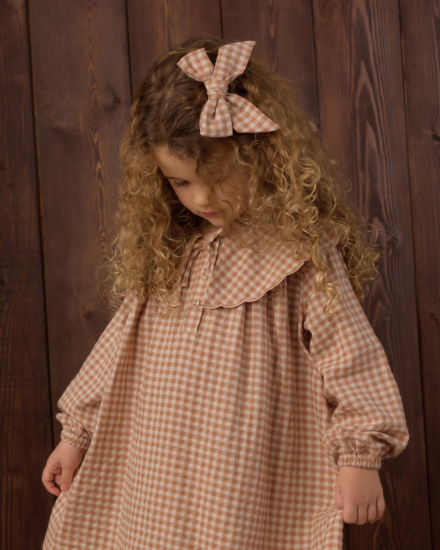 A young girl with long curly light brown hair wearing a brown and beige gingham dress with a large collar and matching bow headband, standing against a dark wood plank background.
