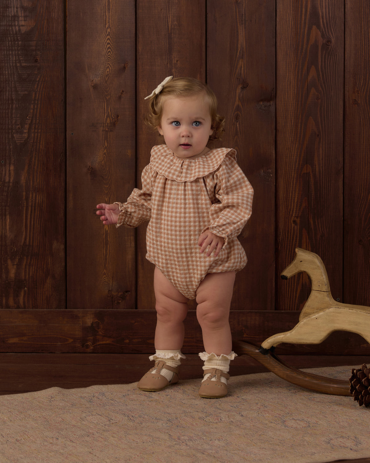 A baby girl wearing a brown and beige gingham long-sleeve romper with a ruffled collar, standing in front of a dark wood plank wall next to a wooden rocking horse.