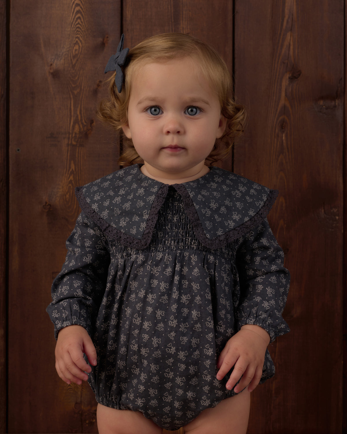 A toddler girl with light brown hair and blue eyes wearing a dark gray floral print romper with a wide smocked collar, standing against a dark wood plank background.