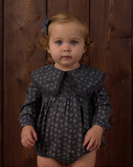 A toddler girl with light brown hair and blue eyes wearing a dark gray floral print romper with a wide smocked collar, standing against a dark wood plank background.