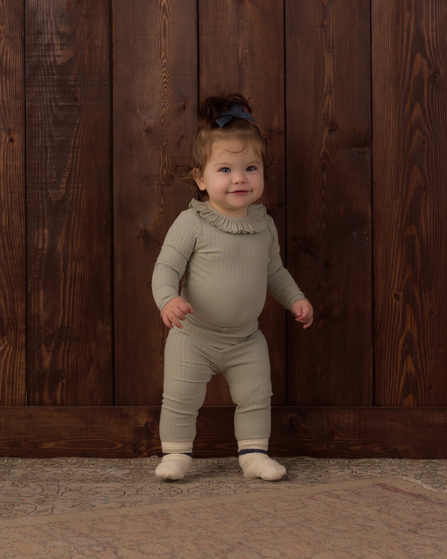 A toddler standing against a dark wood plank wall, wearing a ribbed long-sleeve one-piece jumpsuit in a muted sage green with a ruffled neckline and matching foot coverings.