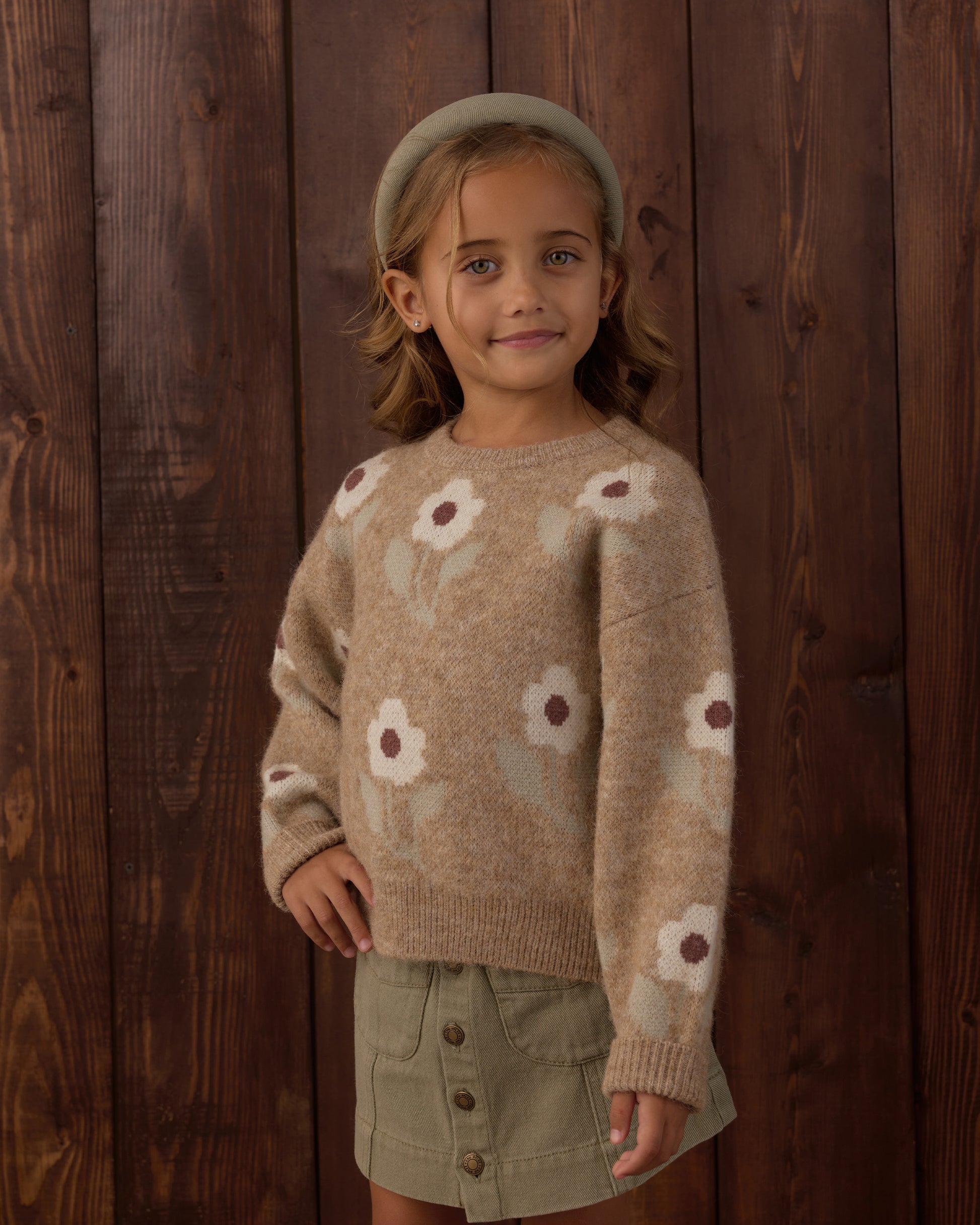 A young girl wearing a beige knit sweater with a white and brown daisy pattern, an olive green skirt, and a matching olive green beret, standing against a dark wood plank background.