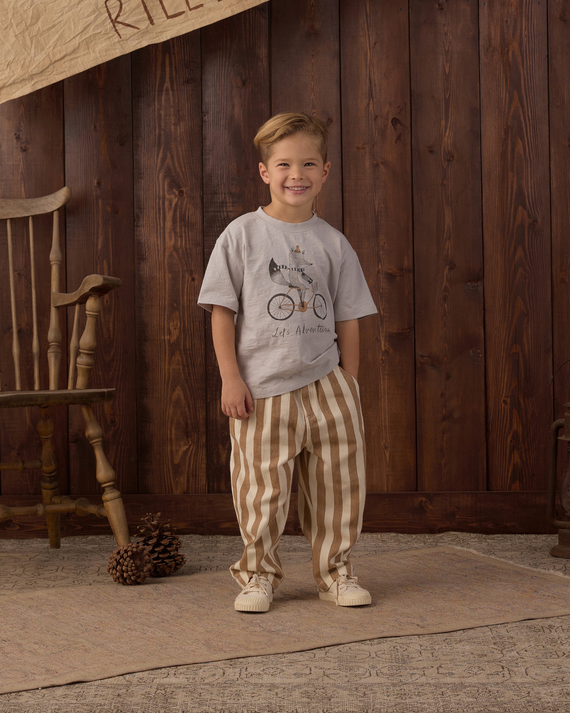 A young boy stands in front of a dark wood plank wall wearing a light gray graphic t-shirt with a bicycle print and beige and brown vertically striped pants.