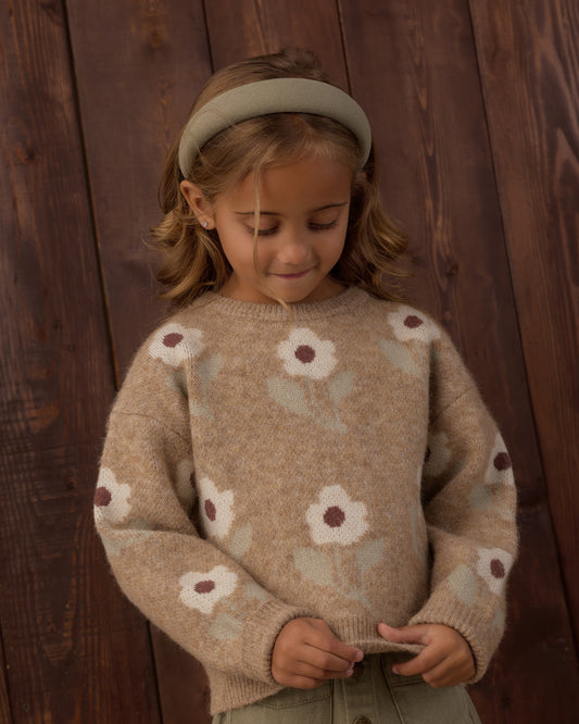 A young girl wearing a beige knit sweater with a floral pattern, a matching beige headband, and olive green pants, standing against a dark wood plank background.