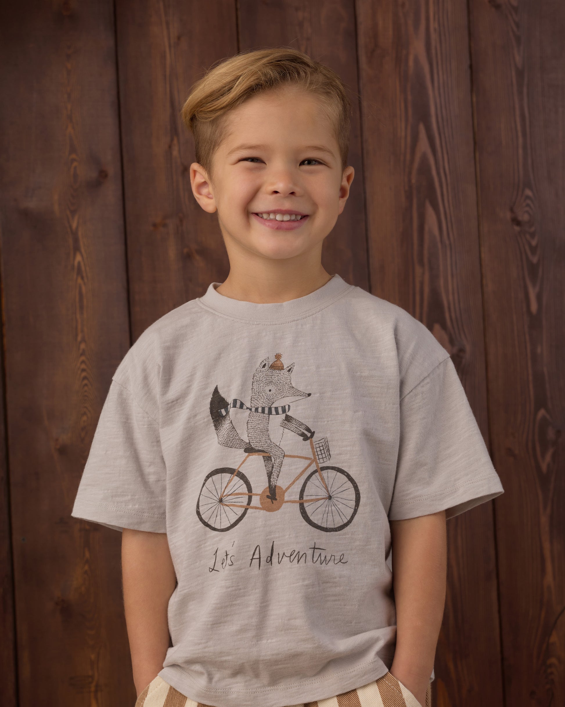 A young boy smiling while wearing a light gray heathered t-shirt with a graphic of a fox riding a bicycle and the text 'It's Adventure'. He is standing in front of a dark brown wooden plank wall.