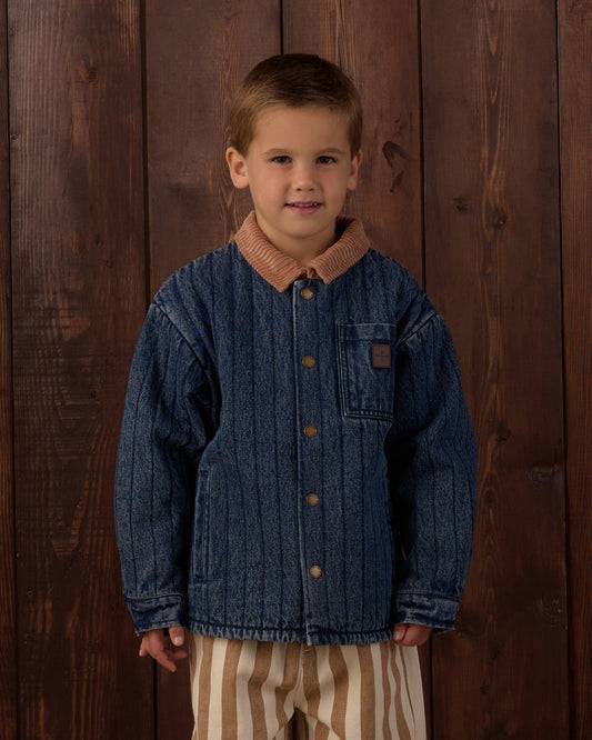A young boy wearing a blue denim jacket with a brown corduroy collar and striped pants, standing against a dark brown wooden plank background.