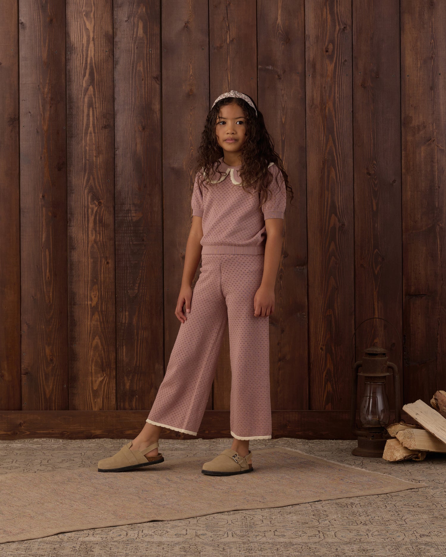 A young girl stands in front of a rustic dark wood plank wall wearing a dusty rose pink two-piece knit set consisting of a short-sleeve top with a white collar and matching wide-leg cropped pants. She is wearing beige slip-on shoes and a white headband.